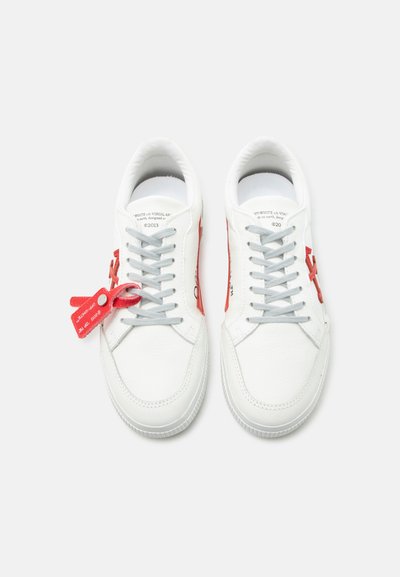 Ζευγάρι λευκών δερμάτινων αθλητικών παπουτσιών με γκρι κορδόνια, κόκκινες λεπτομέρειες στα πλαϊνά και κόκκινο δέσιμο φερμουάρ στη δεξιά κορδόνι του αριστερού παπουτσιού.