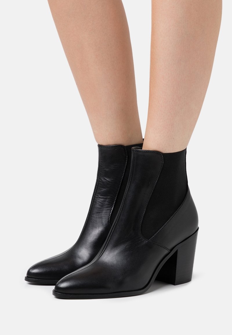 Bottines noires en cuir avec bouts pointus et talons blocs portées sur jambes nues sur fond blanc.