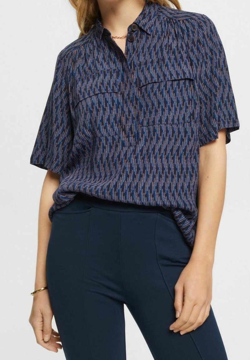 Blusa blu navy a maniche corte con un motivo astratto multicolore, due tasche frontali e colletto classico. Tessuto leggero e morbido.