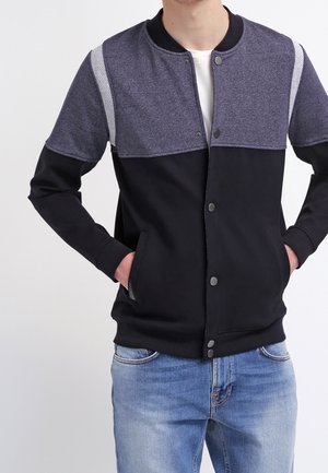 Mann, der eine schwarz-graue Jacke mit Knöpfen und Taschen trägt, kombiniert mit hellblauen Jeans, stehend vor einem einfarbigen hellen Hintergrund.