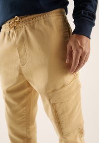 Beżowe spodnie cargo z elastycznym pasem, regulowanym sznurkiem, boczną kieszenią i gładką fakturą materiału, częściowo widoczna dłoń oparta na udzie.
