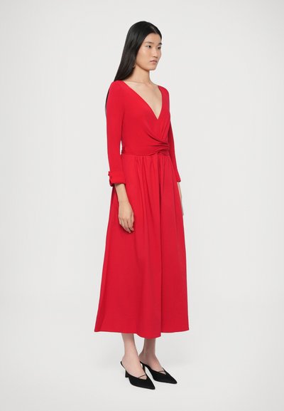Vörös átlapolt ruha mély V-nyakkivágással, hosszú ujjakkal és összegyűjtött derékkal. Sima textúrájú, térd alá érő szegély fekete, minimalista mules cipővel párosítva.