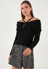 Žena u crnom topu bez naramenica od rebraste tkanine s dugmadi u obliku srca i crno-bijelim šarenim šortsevima, pozira protiv jednostavne pozadine.