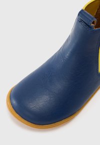 Botín de cuero azul liso hasta el tobillo con fuelle elástico amarillo y suela de goma marrón claro sobre un fondo blanco.