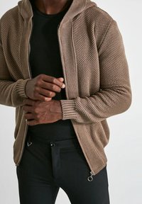 Felpa con zip lavorata a maglia color beige, dal finish testurizzato e con polsini a costine, indossata sopra una maglietta nera. I pantaloni neri presentano dettagli con zip.