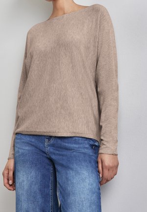 Personne portant un pull à manches longues beige et un jean bleu, debout contre un mur gris clair uni, les mains détendues le long du corps.