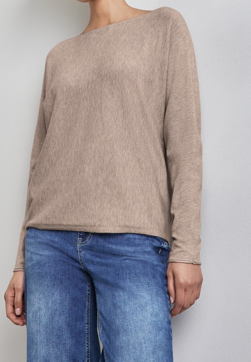 Personne portant un pull à manches longues beige et un jean bleu, debout contre un mur gris clair uni, les mains détendues le long du corps.
