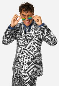 Traje a rayas en blanco y negro con un diseño de estrellas y rayas; el modelo sostiene unas gafas de sol en forma de estrella de arcoíris, vistiendo una camisa y corbata azules.