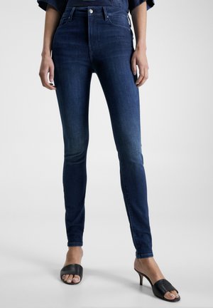 Vrouw draagt donkerblauwe skinny jeans en zwarte open-toe sandalen met hak, staand tegen een effen lichtgrijze achtergrond.