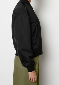 Chaqueta bomber negra con acabado liso y brillante, cuello y puños acanalados, y bolsillo lateral, llevada sobre una falda verde.