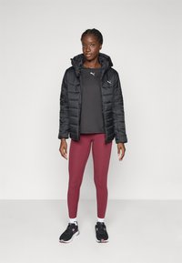 Sort dunjakke med hætte, grå langærmet skjorte, burgundy leggings og sorte sneakers med lyserøde detaljer, stående på en ensfarvet baggrund.