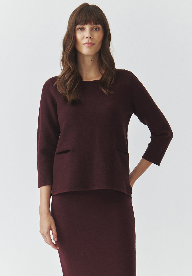Maglione bordeaux con maniche a tre quarti e tasche frontali, realizzato in materiale morbido. Abbinato a una gonna coordinata. Design semplice.