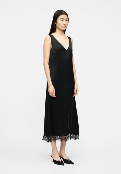 Γυναίκα που στέκεται, φορώντας μαύρο μίντι φόρεμα χωρίς μαν ίκια με V-λαιμόκοψη και δαντελένιο τελείωμα, συνδυασμένο με μαύρα μυτερά παπούτσια με χαμηλό τακούνι.