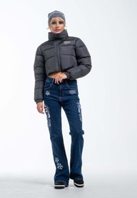 Schwarze, kürzere Daunenjacke mit hohem Kragen, kombiniert mit blauen Jeans im Schlag, die weiße Blumenstickerei und ein dezentes Logodesign aufweisen.