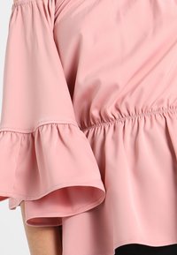 Haut rose à épaule dénudée avec des manches évasées, présentant un élastique froncé à la taille et une texture de tissu lisse et léger.