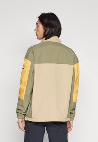 Giacca con un corpo beige, parte superiore verde e maniche gialle. Presenta polsini elastici, colletto alto e un dettaglio con logo lungo il lato.