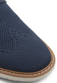 Zapatilla de malla de color azul oscuro con una superficie texturizada, acentos marrones y una suela blanca. Presenta costuras decorativas y un diseño de puntera redondeada.