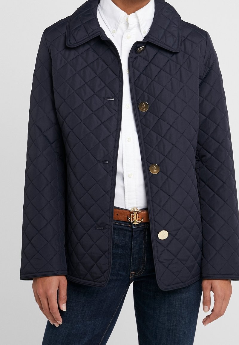 Vadderad marinblå jacka med klassisk krage, med guldknappar och sidofickor, i kombination med en vit skjorta och ett brunt bälte.