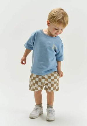 Fiatal gyermek lefelé néz, kék inget, barna-fehér kockás rövidnadrágot, szürke zoknit és fehér tornacipőt visel egyszínű háttéren.