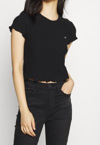 Woman wearing a textured black short-sleeve cropped top and black high-waisted jeans, with one hand in pocket against a white background.