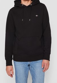 Sudadera negra con un bolsillo frontal, cordones y un pequeño logo bordado en el pecho. Hecha de una tela suave, presenta puños y dobladillo acanelados.