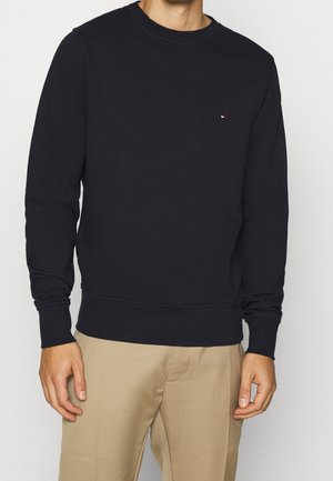 Muž v jednoduchém černém svetru s dlouhým rukávem a kulatým výstřihem a v béžových kalhotách, stojící před bílým pozadím.