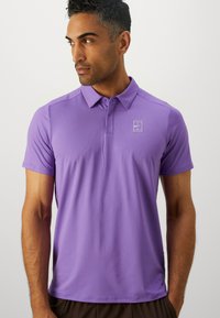 Muškarac nosi ljubičastu Nike polo majicu s ovratnikom, stojeći protiv jednostavne svijetle pozadine, gledajući ulijevo.