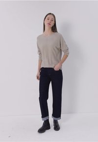 Kobieta w beżowej bluzce z długim rękawem, ciemnych dżinsach z mankietami i czarnych botkach na kostkę stoi na tle białego tła.