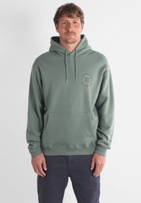 Hoodie vert en tissu doux, doté d'une poche avant, de poignets côtelés et d'un imprimé de logo circulaire sur la poitrine. Le modèle est debout.