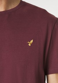 Burgunderfarbene Baumwoll-T-Shirt mit kleinem, goldenen, gestickten Vogel-Logo auf der Brust. Kurze Ärmel und runder Ausschnitt. Weiche Textur.