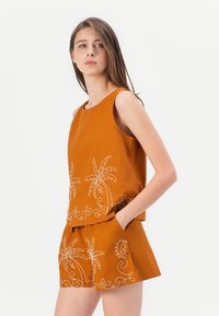Oranges ärmelloses Oberteil mit weißer bestickter Palmen-Design, kombiniert mit passender Shorts, die ähnliche Stickerei aufweisen, beide aus einem weichen Stoff.