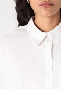 Camisa branca de botão com colarinho clássico, apresentando uma textura suave e um leve brilho. Inclui um bolso frontal e detalhes em botões de pérola.
