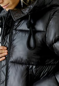 Zwarte pufferjack met een gestructureerde afwerking, voorzien van een cinch-trekkoordkap, een ritszak en subtiele branding op de stof.