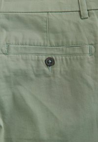 Tela verde claro con una textura suave. Presenta un bolsillo trasero con un cierre de botón oscuro y un detalle de costura sutil.