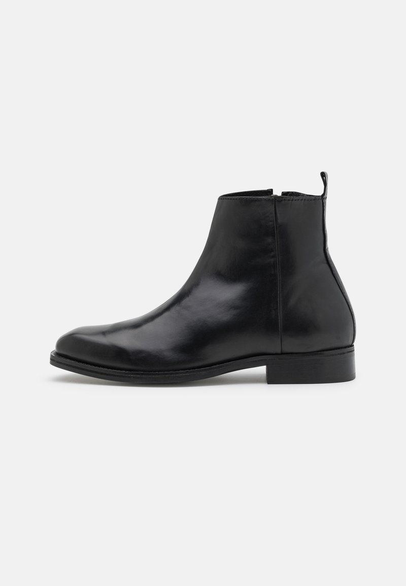 Bottine en cuir noir lisse à talon bas, avec fermeture éclair latérale et languette de tirage sur fond uni gris clair.