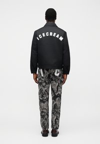 Μαύρο μπουφάν με λευκό κείμενο «ICECREAM» στην πλάτη, συνδυασμένο με γκρι παντελόνι με μοτίβα. Η εμφάνιση έχει άνετη εφαρμογή και ομαλή υφή.