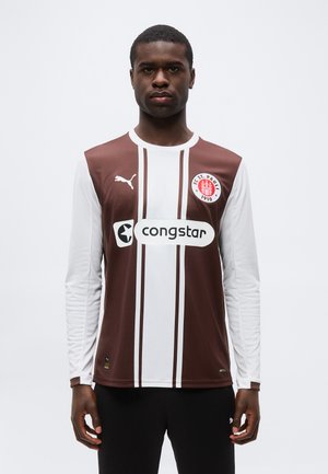 Mand iført FC St. Pauli brun og hvid stribet langærmet fodboldtrøje med "congstar" sponsorl ogo, stående mod ensfarvet baggrund.