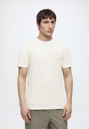 Ung mand iført en enkel lysebeige t-shirt med korte ærmer og et subtilt rundt logo på venstre bryst side, stående foran en hvid baggrund.