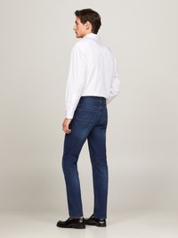 Jeans blu scuri con vestibilità slim, dotati di tasche posteriori cucite. Abbinati a una camicia bianca a maniche lunghe e scarpe nere.