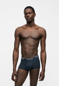 Λεπτός μυώδης άνδρας που φοράει μπλε σκούρο boxer Calvin Klein στέκεται μπροστά σε λευκό φόντο με ουδέτερη έκφραση.