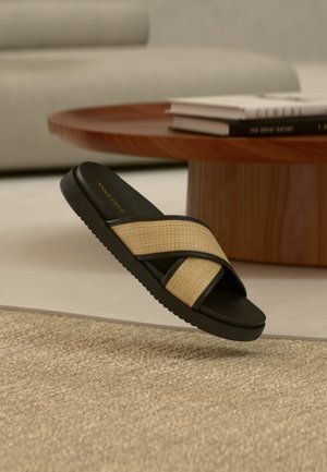 Sandale noire à enfiler avec des lanières croisées tressées de couleur beige, dotée d'une semelle en caoutchouc souple et d'une finition texturée. Nom de la marque "ANNA FIELD" en doré.