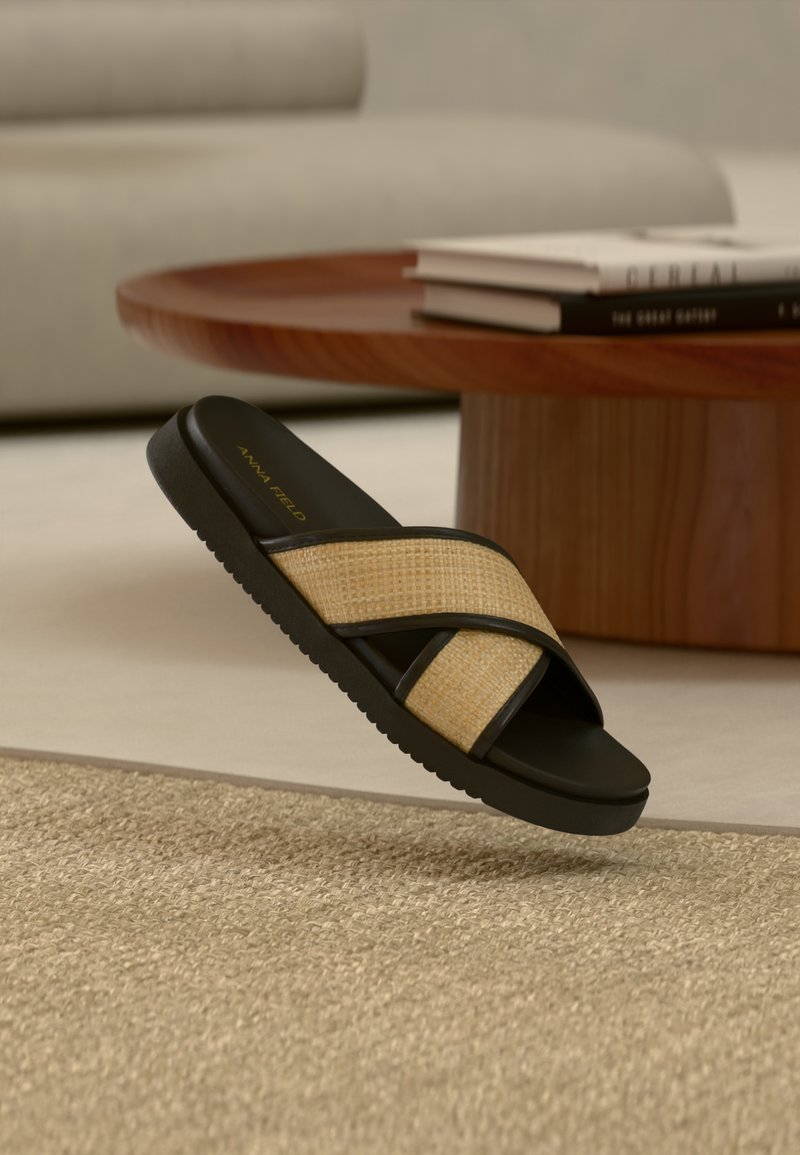 Sandale noire à enfiler avec des lanières croisées tressées de couleur beige, dotée d'une semelle en caoutchouc souple et d'une finition texturée. Nom de la marque "ANNA FIELD" en doré.