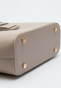 Base della borsa a tracolla in pelle beige con quattro piedi in metallo dorato, texture liscia e bordi cuciti. Presenta un dettaglio con pattina superiore.