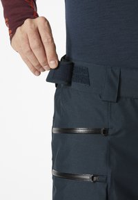 Tmavě modré kalhoty mají suchý zip na pásu a tři kapsy na zip s černými kovovými doplňky. Materiál má texturovaný vzhled a úzký střih.