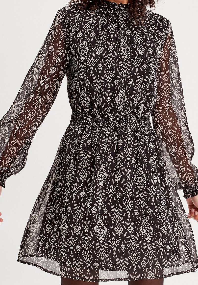 Robe noire à motif floral avec manches longues en voile transparent, taille élastique, tissu léger, encolure froncée, longueur jusqu'aux genoux.