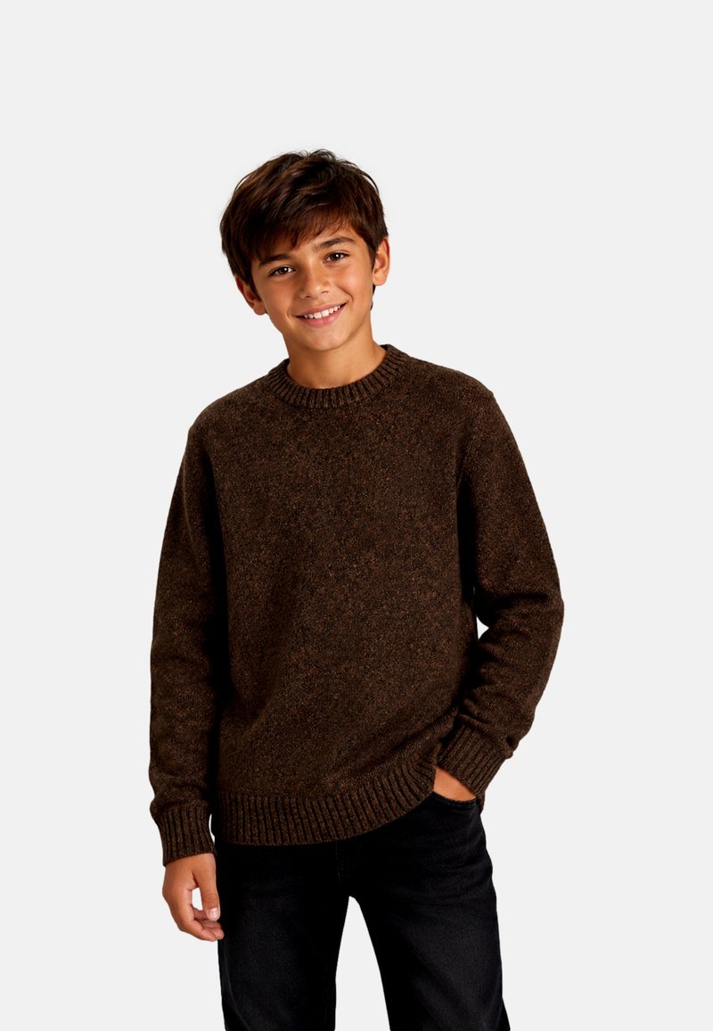 Garçon aux cheveux bruns portant un pull en laine marron foncé et un pantalon noir, souriant avec une main dans sa poche devant un fond uni.