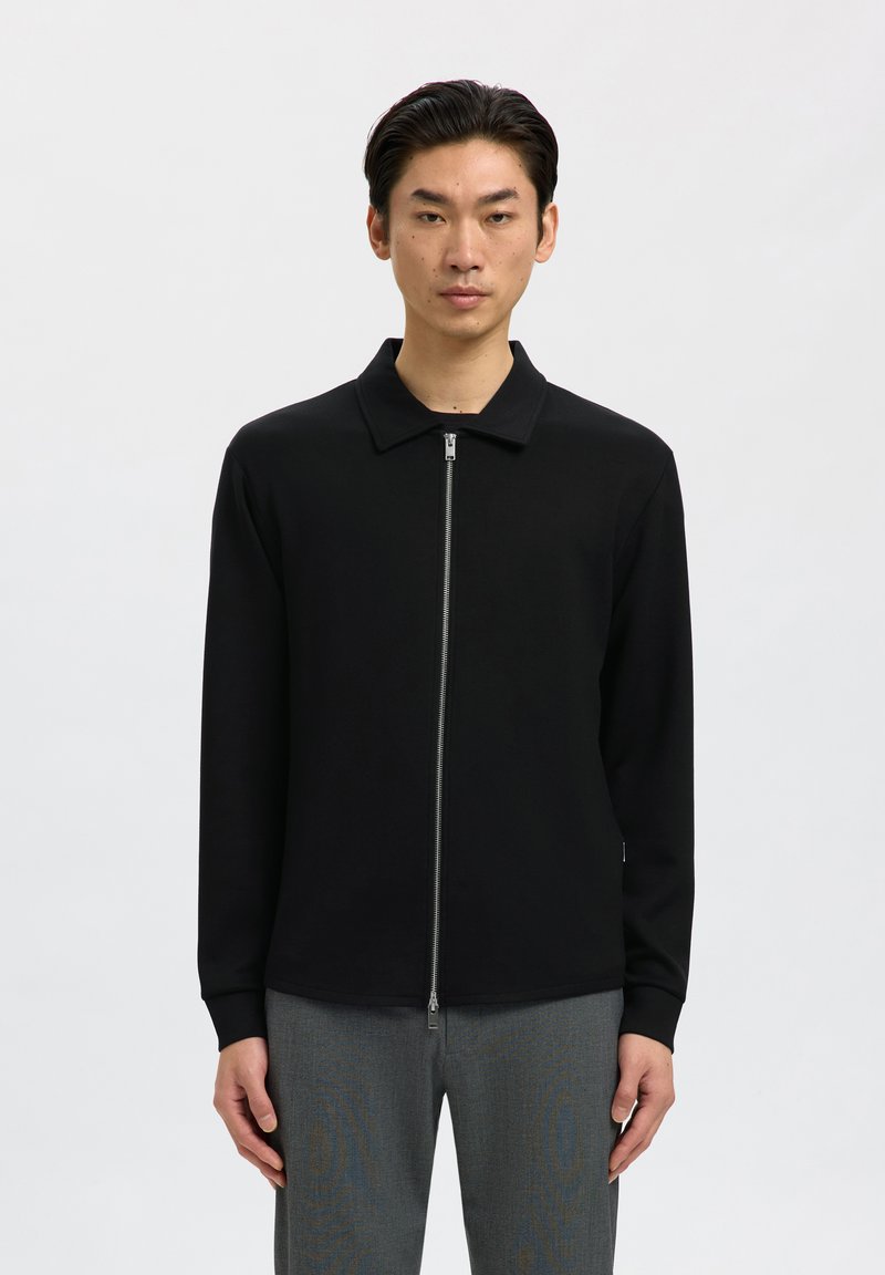 Homme portant une veste noire zippée avec col et un pantalon gris, debout devant un fond clair uni.