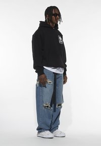 Felpa nera con cappuccio e logo frontale, abbinata a jeans strappati blu chiaro e sneakers bianche. Gli occhiali da sole completano il look casual.