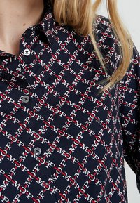 Camicia blu navy con bottone, caratterizzata da un motivo ripetuto di logo rosso e bianco. Il tessuto ha una texture liscia, con un colletto classico e una chiusura a singolo bottone.