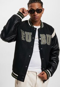 Sort varsity-jakke med creme- og olivengrønne detaljer, læderærmer, oversized bogstaver og ribbede manchetter. Har snap-knap lukning.